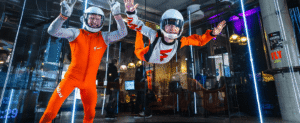 A child and an adult flyer in bright orange Fööni flight suits experiencing indoor skydiving together inside the vertical wind tunnel.