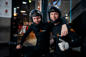 Two happy men in black indoor skydiving suits and helmets smiling and giving a thumbs up at the Fööni flight center.