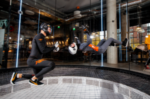 A professional Fööni instructor provides hands-on coaching to a beginner student practicing stable body position on the flight chamber's safety net.